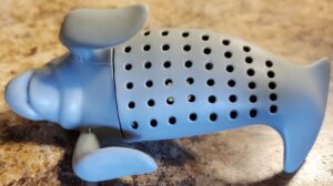 A tea-infuser shaped like a manatee. The manatee is a light grey-blue color, with fins extending away and down from its body in a 90 degree angle to facilitate it sitting on cup rims. In the main section of the body there are small holes in the plastic for draining tea leaves and there is a seam between the head and body to open the infuser to add or remove tea. It sits on a brown and tan faux stone kitchen laminate. 