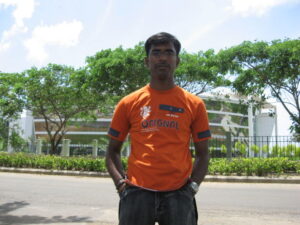 Photograph of a presumed South-Asian man. He stands outdoors in front of a road near some greenery and wears an orange shirt with his hands in his pockets. 