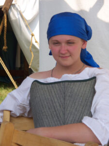 A young white woman sitting in a wooden chair in front of a white tent. She wears a blue headscarf, white peasant blouse, and grey stays.