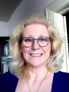 A Photo of a middle-aged blonde woman. She is white with glasses, a necklace, and a blue top. She is smiling and appears friendly.